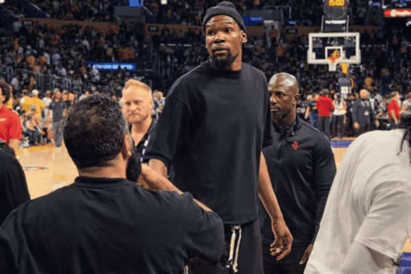 Basketball player in a black shirt standing on a court, surrounded by teammates and staff with a crowded arena in the background.