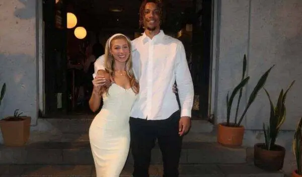Couple posing arm-in-arm outside a doorway at night; woman in a white bodycon dress, man in a white button-down shirt and black pants.