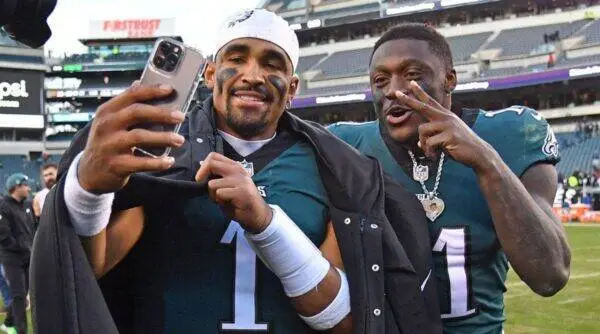 Two football players in teal jerseys take a postgame selfie on the field, one flashing a peace sign toward the camera while smiling.