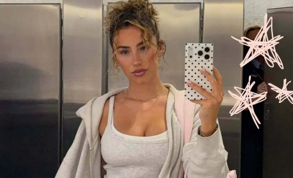 Selfie of a woman in a metallic elevator wearing a white tank top and light gray hoodie, holding a white phone with a black polka-dot case.