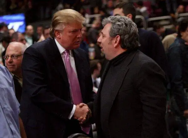 Two men in dark jackets shake hands in a crowded event, one with a pink tie and light hair.