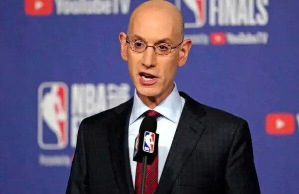 Bald man in a suit speaks at a podium with an NBA logo microphone at a blue press backdrop
