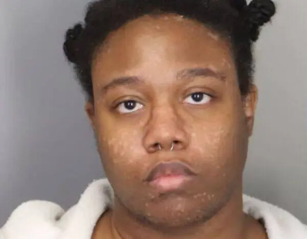 Mugshot-style portrait of a young person facing the camera, wearing a light robe, with a septum piercing.