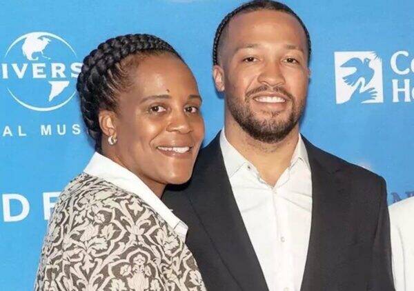 Smiling couple posing at a blue event backdrop; woman in a patterned blazer with braided hair, man in a black suit and white shirt.