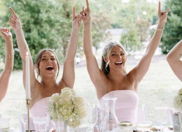 Three bridesmaids in pink strapless dresses cheer with arms raised at an outdoor wedding reception fall back greenery behind them.