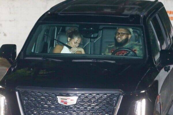 Man driving a black Cadillac SUV with a bearded man wearing glasses in the driver’s seat and a woman in the passenger seat resting her head on her hand.
