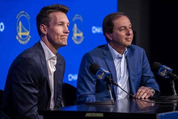 Two men in suits at a press conference, microphones in front, blue backdrop with logos behind them in a panel setting.