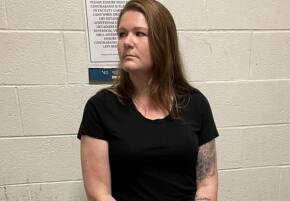 Woman with shoulder-length brown hair in a black t-shirt stands against a white cinder block wall, left forearm tattoo visible.