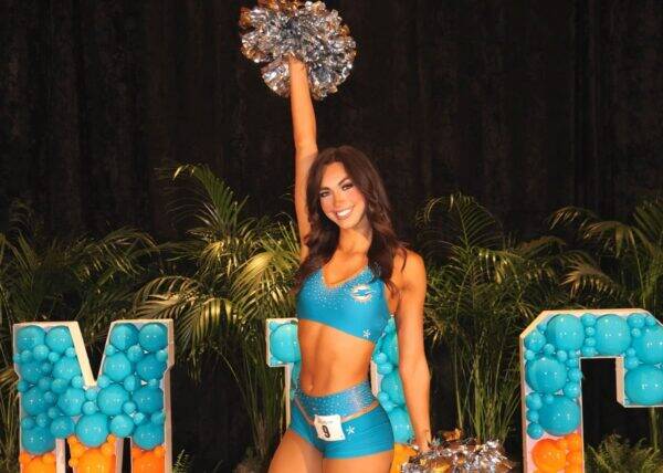 Cheerleader in a turquoise uniform raises a silver pom-pom on stage, smiling, with teal balloon letters in the background.