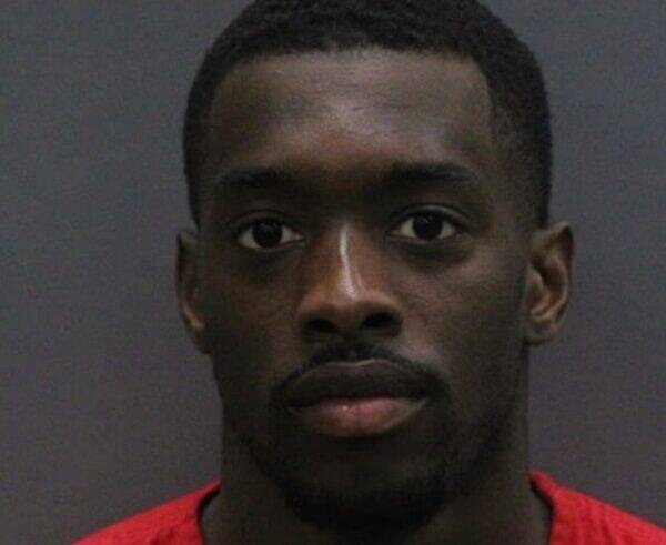 Mugshot of a young Black man with short hair, wearing a red shirt, facing the camera against a gray background.