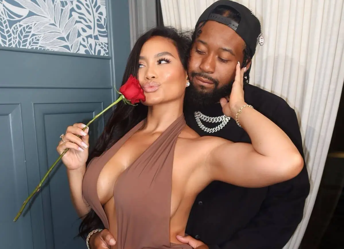 Woman in a taupe halter dress poses with a red rose near her lips while a man hugs her from behind.