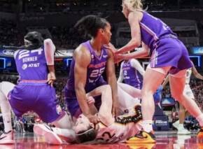 Female basketball players in purple jerseys attend to a teammate lying on the court during a game in progress to the left.