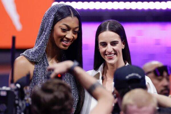 Two women pose for photographers at an event; one wears a sparkling hooded outfit, the other a white shirt, with fans and cameras in the foreground.