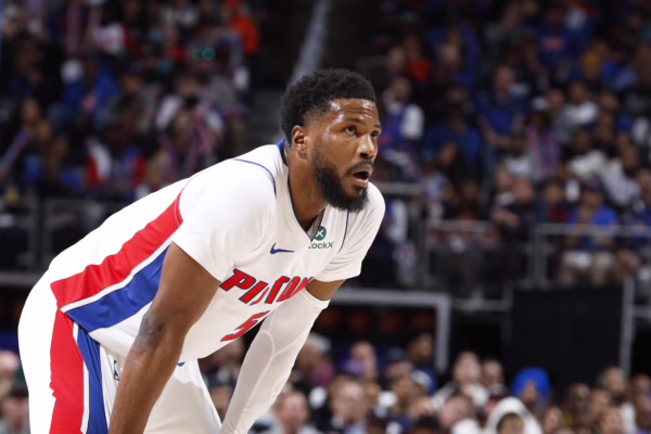 Black male basketball player in a white Pistons jersey with red and blue stripes, bent forward on the court as a crowded arena watches the game.