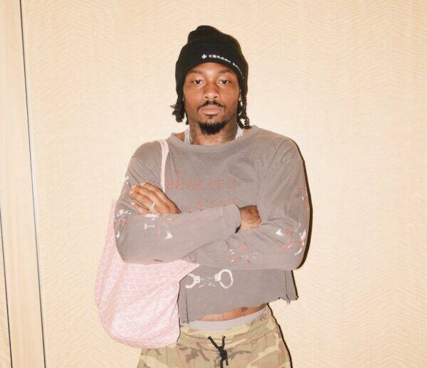 Young man with a black beanie and shoulder bag stands with arms crossed, wearing a paint-splattered gray shirt and camo pants in a neutral background.