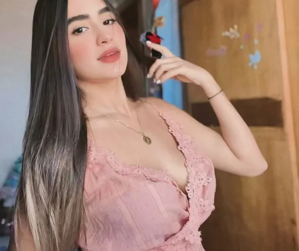 Young woman with long dark hair wearing a pink lace top, posing with her hand near her face indoors.