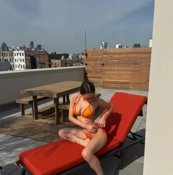 Woman in an orange bikini reclining on a red lounge chair on a rooftop terrace with a wooden privacy screen and city skyline in the distance.
