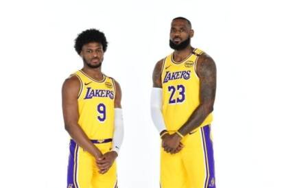 Two Lakers basketball players in yellow uniforms pose together, wearing numbers 9 and 23 against a white background with white arm sleeves on both arms, left and right respectively.