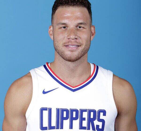 Portrait of a smiling male basketball player in a white Clippers jersey against a blue background, showing his upper torso and shoulders.