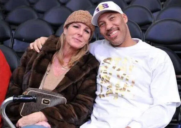 Smiling couple sitting close together in stadium seating; woman in fur coat and brown knit hat, man in white shirt and cap with gold lettering.