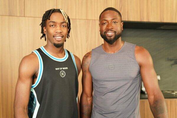Two smiling men in sleeveless athletic tops posing side by side in a wood-paneled gym area.