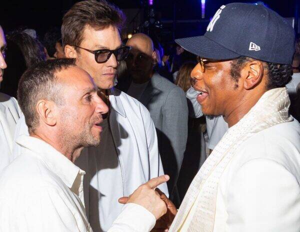 Three men in white clothing converse at a crowded event; the man on the left points at the man wearing a navy NY Yankees cap.
