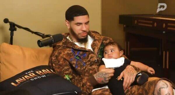 Bearded man in a camo hoodie sits on a couch holding a baby, with a microphone in front for a podcast.