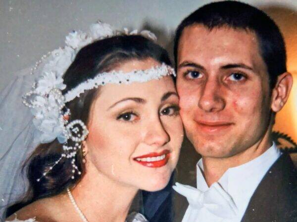 Bride and groom pose for a close-up wedding photo, she wears a lace headband and veil, he in a tuxedo with a white bow tie, both smiling.