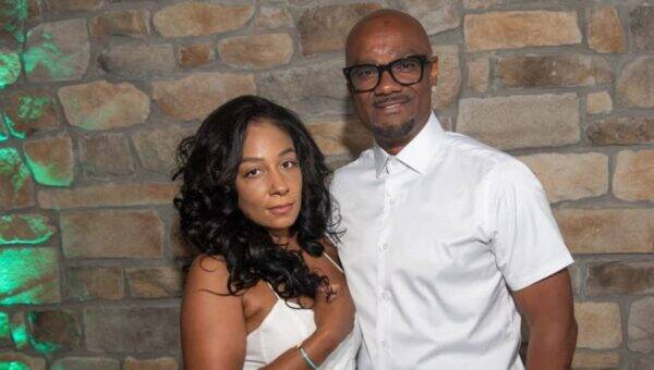 Couple posing together indoors against a stone wall, both in white outfits and smiling at the camera.