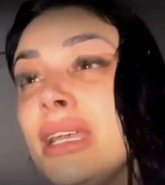 Close-up of a woman's face with dark hair, mouth open mid-speech or expression, makeup visible.