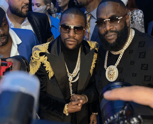 Two men in flashy attire pose for photos at a crowded event: one with a black and gold embroidered jacket and layered gold chains, the other in a black Fendi-knit top with a large gold medallion; several photographers and people surround them.