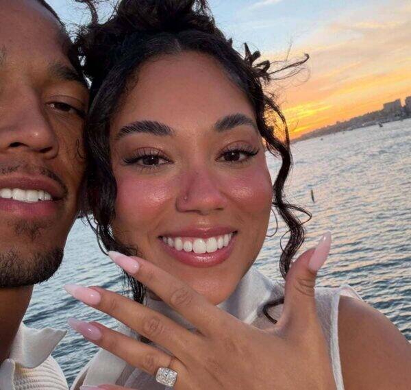 Couple taking a sunset selfie by the water; woman in front shows off a diamond ring on her finger and pink nails, man beside her smiling. The ocean and city skyline are in the distance.