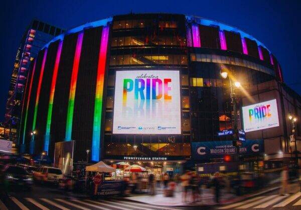 Night view of Madison Square Garden with rainbow Pride lights and a large screen reading 'PRIDE' while crowds gather outside.