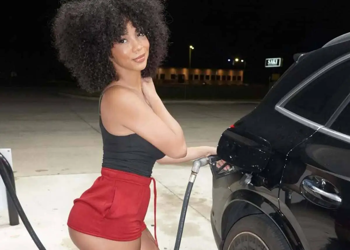 Woman in a black tank top and red shorts refuels a black car at a gas station at night.