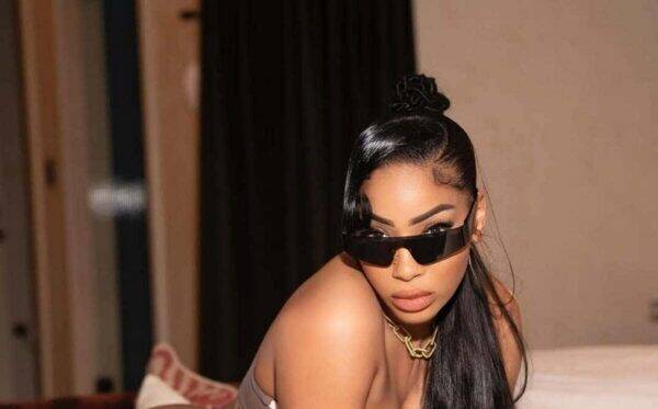 Woman with long black hair wearing oversized black sunglasses and a chunky gold necklace, indoors on a bed.