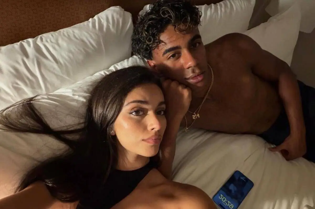 Couple lying on a bed together; woman in front taking a selfie while a shirtless man rests beside her wearing gold chains.