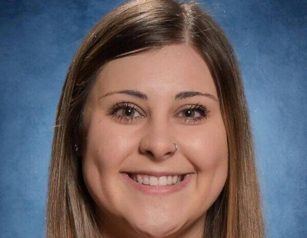 Close-up portrait of a smiling woman with brown hair and a nose ring against a blue studio backdrop.