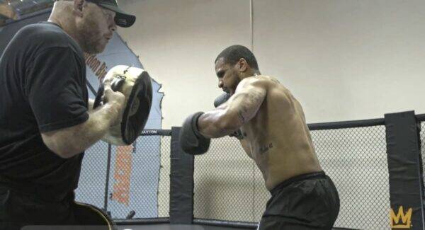 Boxer practicing punches with a trainer holding focus mitts inside a training ring in a gym cage.