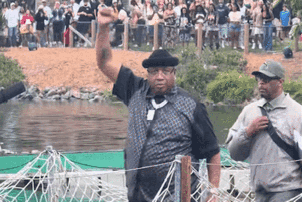 Man in a black hat and checkered vest raises his right fist at a waterfront event, crowd behind him.