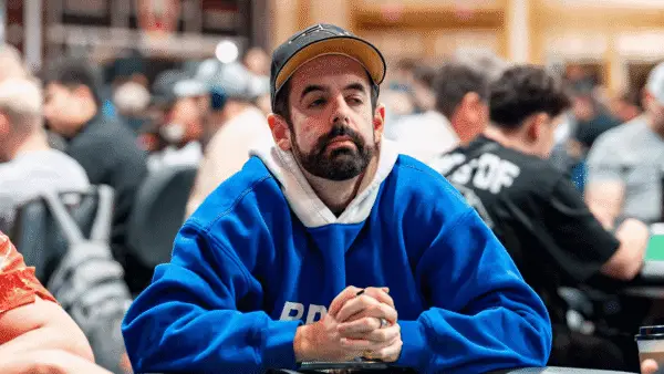 Man with a beard in a blue hoodie and brown cap sits at a crowded table, hands clasped, looking thoughtful at an indoor event.