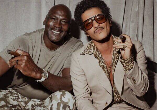 Two men posing for a fashion photo, one in a beige tee with camo pants and the other in a tan suit with leopard-print shirt and sunglasses, both holding cigars and smiling.