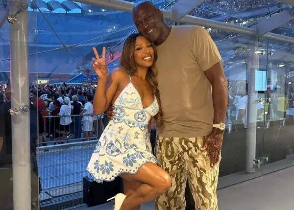 Happy couple posing for a photo at a crowded indoor event; woman in a blue-and-white floral dress makes a peace sign beside a tall man in a tan shirt and camo shorts.