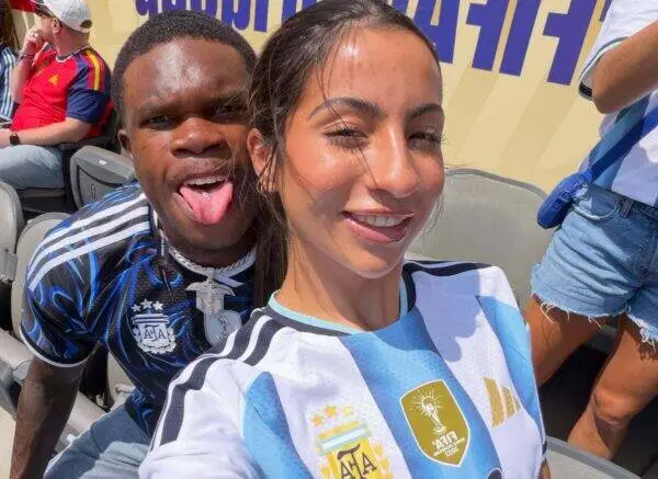 Two friends pose for a selfie in stadium seats; woman in an Argentina football jersey smiles, man behind her sticks out his tongue.