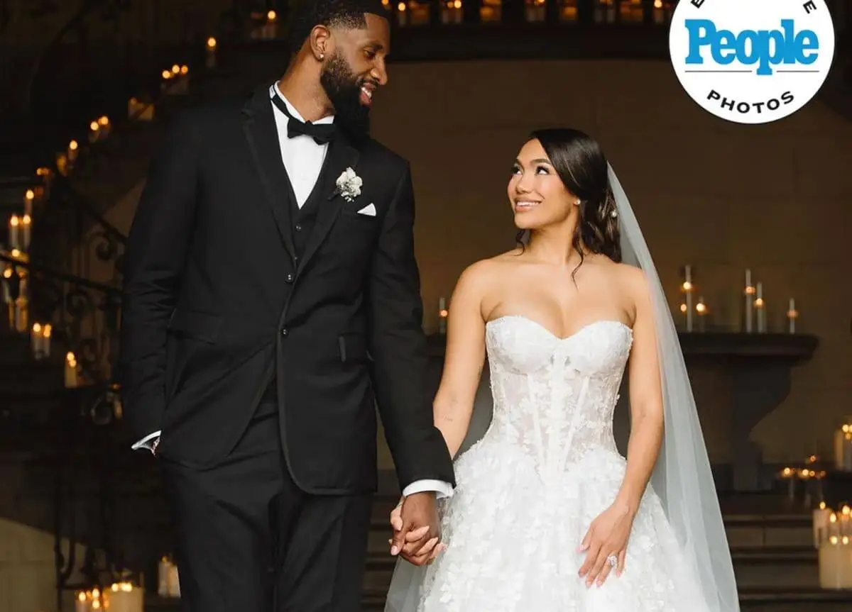 Bride in a white lace strapless gown and veil, holding hands with a groom in a black tuxedo on a candlelit staircase.