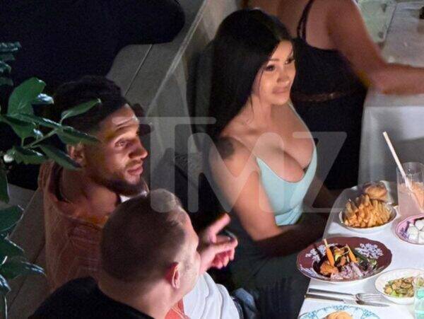 Group of people dining at a table with plates of food, including a woman in a light blue dress seated beside a bearded man.