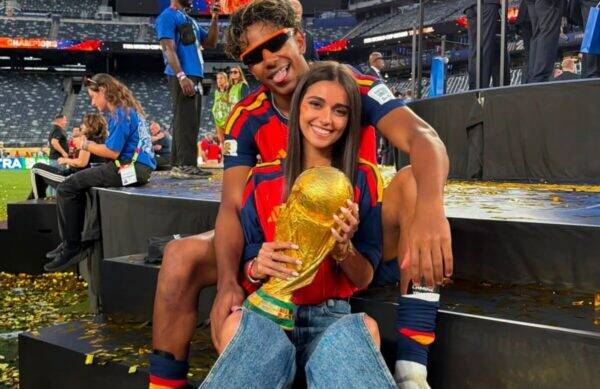 Two young athletes in red-blue jerseys sit on stadium steps, smiling as the woman holds a large gold trophy amid confetti on the ground.
