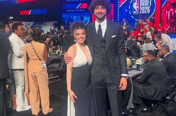 Tall man in a dark pinstripe suit and tie stands with a smiling woman in a white halter top and black skirt at a crowded formal event, with bright NBA Draft 2026 screens in the background.