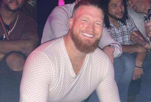 Smiling bearded man in a white mesh shirt posing for a group photo at a nightclub, with friends in the background.