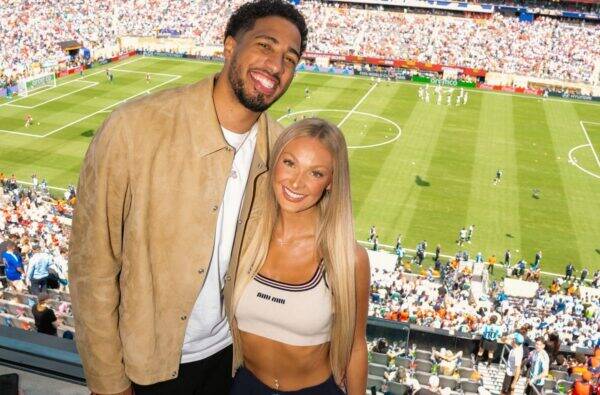 Smiling couple posing for a photo in a soccer stadium with a green pitch and cheering spectators in the stands.