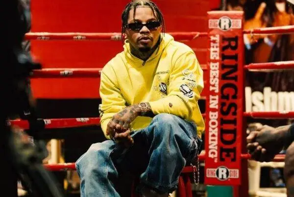 Man wearing a yellow hoodie and sunglasses sits in a boxing ring, arms folded, with red ropes and a Ringside banner nearby.
